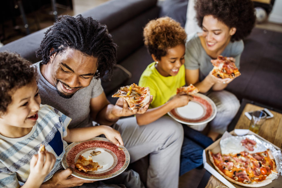 Happy parents and their small kids eating pizza in the living room.