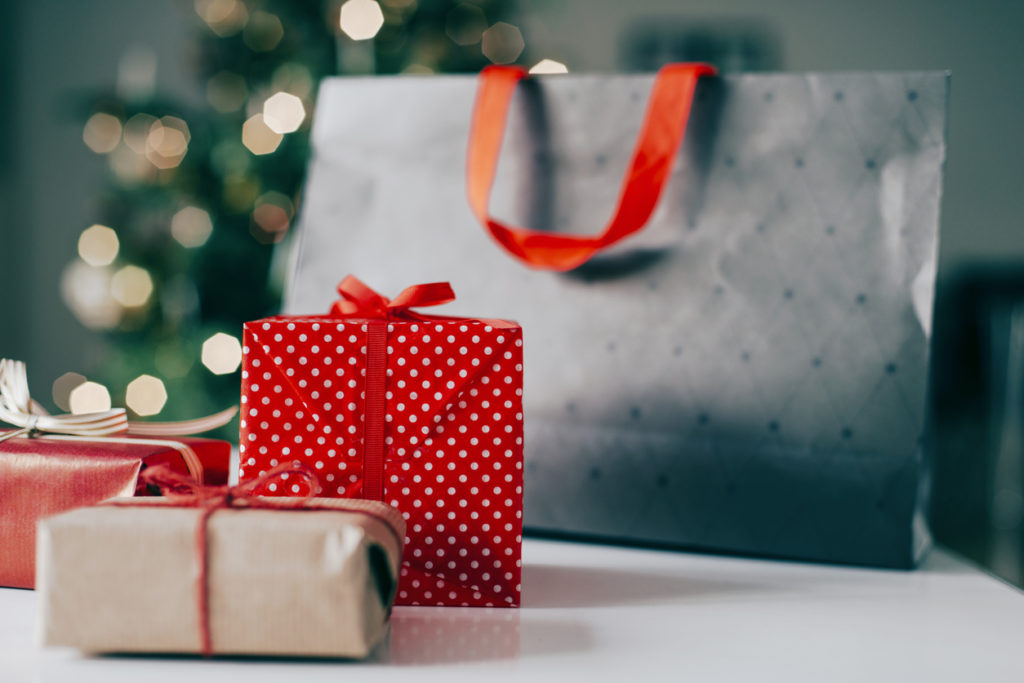 Several Christmas presents wrapped in different wrapping paper has been placed on a table next to a shopping bag. Blurred out christmas tree in the background.