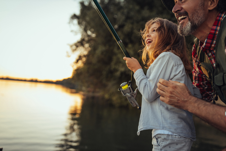 Grandfather showing his granddaughter how to fish while standing on the dock by the river.